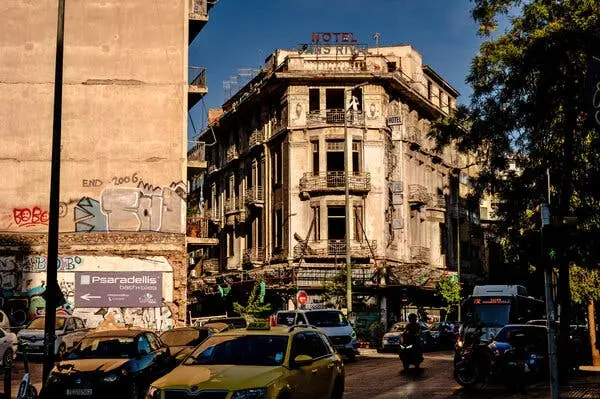 نمایی از خیابان که ساختمانی هتل فرسوده با تابلو قرمز 'Hotel Sans Rival' در بالای آن را نشان میدهد. خودروها و یک موتورسیکلت از کنار دیوار پر از گرافیتی عبور میکنند.