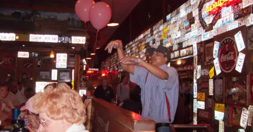 مردی با پیراهن دکمهدار آبی و سفید راهراه، شلوار کشسان قرمز و کلاهی آبی، نان رول را در سالن غذاخوری لامبرت پرتاب میکند.