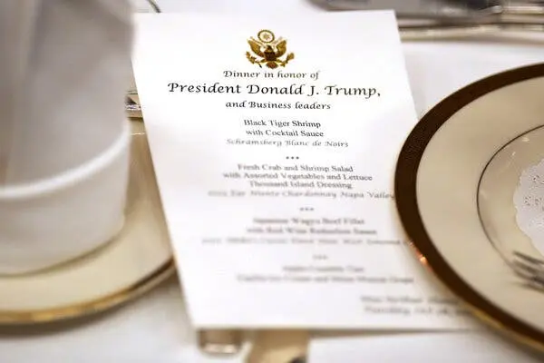 یک منو با عنوان «شام به افتخار پرزیدنت دونالد جی. ترامپ و رهبران کسبوکار» بین بشقابها و کارد و چنگال دیده میشود.