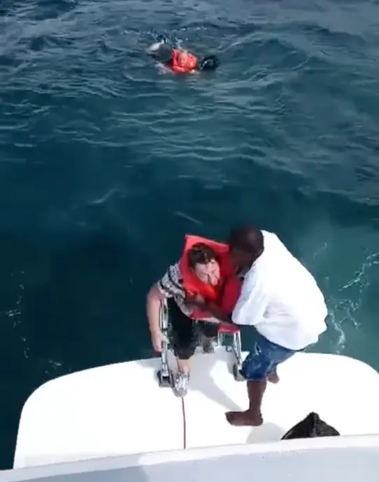 مردی با جلیقه نجات از آب به قایق منتقل می‌شود، در حالی که شخص دیگری با جلیقه نجات در نزدیکی شنا می‌کند.