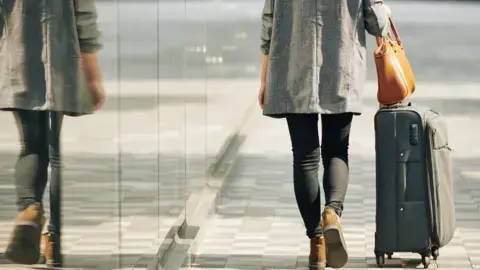 گتی ایمیجز: زنی که پشت به دوربین ایستاده و چمدانی را در خیابان میکشاند.