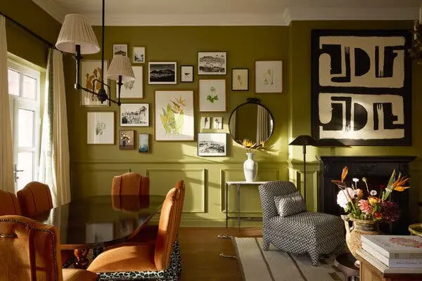 یک سوئیت با دیوارهای سبز و مجموعهای از قابهای عکس. در کنار یک پنجره، میز بیضی شکل با صندلیهای پوشیده به رنگ نارنجی