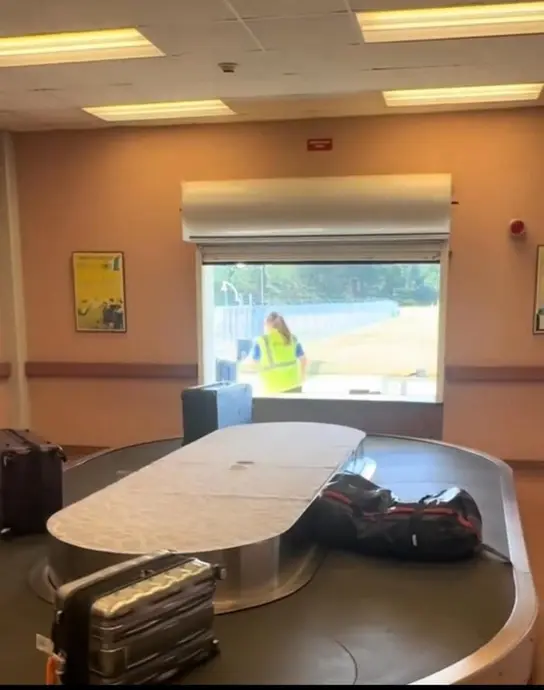 چمدان روی نوار حمل بار در فرودگاه قرار دارد. در پس‌زمینه، کارمند فرودگاه در جلیقه‌ٔ پرنور دیده می‌شود که از پنجره‌ای خارج است.