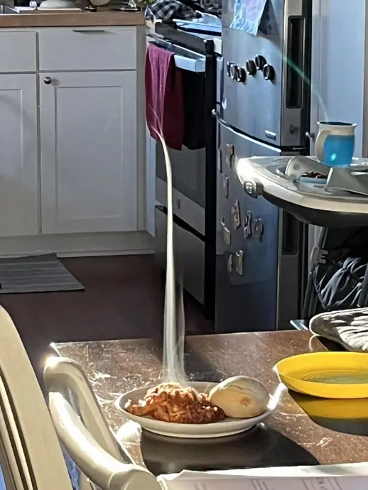 یک بشقاب پاستای بخارآلود با سس و یک رول بر روی میز آشپزخانه. لوازم آشپزخانه در پس‌زمینه قابل مشاهده‌اند.