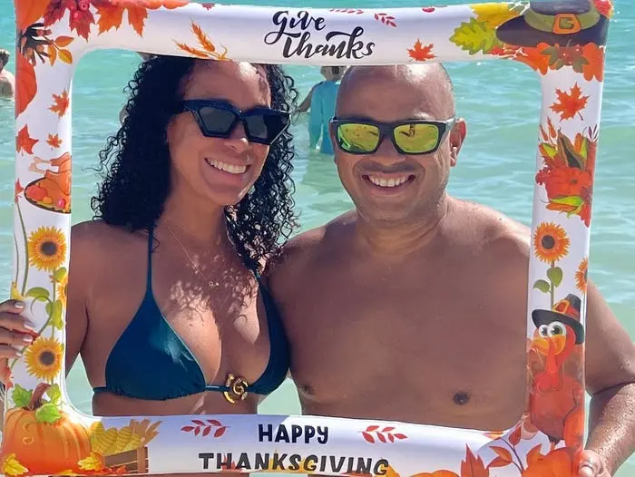 دو نفر در ساحل ایستادهاند و یک قاب با نوشته «از خدا تشکر میکنیم، عید شکرگزاری مبارک» را در دست دارند.