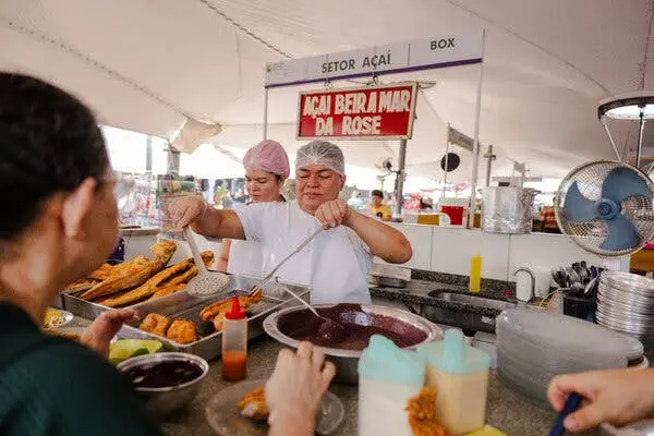 دو نفر غذا را از پیشخوانی که ماهی سرخشده و آساي بنفش دارد، سرو میکنند. یک تابلو قرمز متن "AÇAÍ BEIRA MAR DA ROSE." را نشان میدهد.