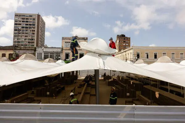 کارگران یک چادر سفید بزرگ و غرفههای چوبی را میسازند. دو نفر بالای چادر با شکل گنبدی آن را تنظیم میکنند، در پسزمینه ساختمانهای شهری قرار دارند.