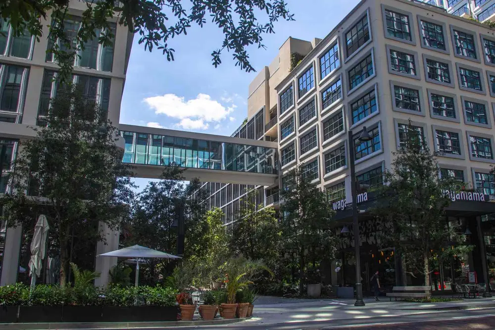 ساختمان‌های مدرن شهری با راه‌پیمایی شیشه‌ای، درختان و صندلی‌های کافه بیرونی در پیش‌زمینه