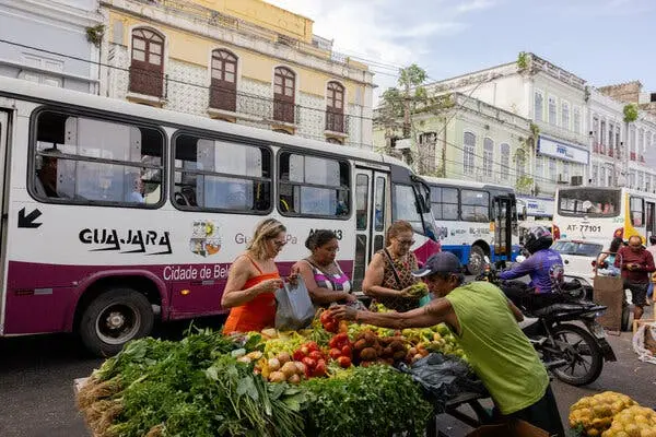 چندین نفر دور یک غرفه بازار خیابانی جمع شده‌اند، محاصره شده توسط سبزیجات و میوه‌های سبز تازه. اتوبوس‌ها در طول خیابانی با ساختمان‌های قدیمی از کنار غرفه می‌گذرند.
