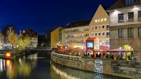 Uwe Niklas/ Tourismus Nuernberg بازار کریسمس نورنبرگ یک "روستای Feuerzangenbowle" کنار رودخانه دارد که کلبه‌ها شراب گرم مشعلی معروف شهر را سرو می‌کنند (اعتبار: Uwe Niklas/ Tourismus Nuernberg)