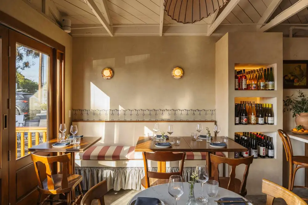دکور داخلی رستورانی با میزها، صندلی‌ها، قفسه‌های شراب و نور طبیعی از پنجره‌ها