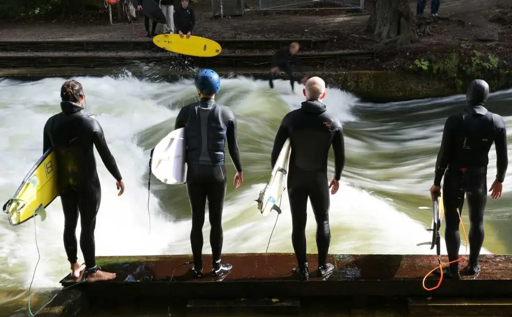 چهار مرد با تخته‌های موج‌سواری خود در کنار موج آیسباخ در «باغ انگلیسی» منتظر هستند.