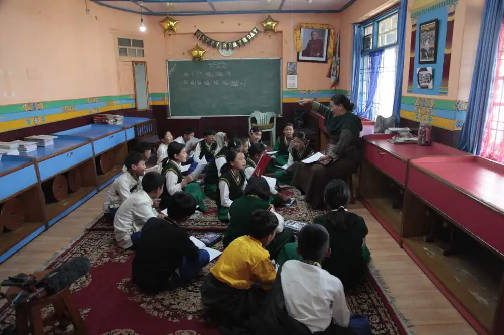 یک معلم موسیقی در حال راهنمایی کلاسی در مدرسه دهکده کودکان تبت. این مدرسه جایگاه ویژه‌ای در میان جامعه تبتی در تبعید در هند دارد. این مرکز شبکه‌ای از مدارس شبانه‌روزی است که به کودکان تبتی زبان، فرهنگ و آیین‌شان را می‌آموزد و توسط خود تبعیدیان به رهبری رهبر معنوی کاریزماتیک‌شان، دالایی لاما، ساخته شده است. اما این مدرسه با چالشی وجودی روبروست: در حال کوچک شدن است زیرا کودک کافی برای پر کردن کلاس‌های پایه‌های پایین‌تر وجود ندارد، که این خود بازتابی از سرنوشت جامعه تبعیدی است. این مسئله در بحبوحه دوره‌ای از عدم قطعیت برای جامعه تبت رخ می‌دهد که خود را برای درگذشت دالایی لاما آماده می‌کنند. عکس از ضیاء حدید، دارامشالا، آوریل ۲۰۲۵