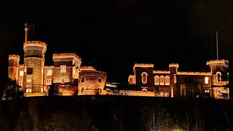 دانکن واکر یک قلعه سنگی باشکوه به طور دراماتیک در نورهای طلایی گرم در برابر آسمان شب سیاه روشن شده است. برجها و باروهای آن با جزئیات معماری تیز برجسته شده و یک silhouette چشمگیر ایجاد کردهاند.