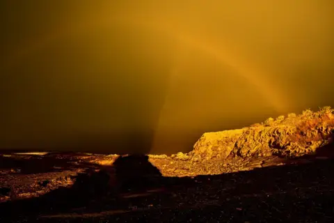 جان کاتبرت یک منظره ناهموار زیر آسمانی زرد-طلایی شدید میدرخشد و سایههای دراماتیک روی زمین میاندازد. یک رنگینکمان کمرنگ از میان نور سوررئال عبور میکند و نرم به افق میپیوندد.