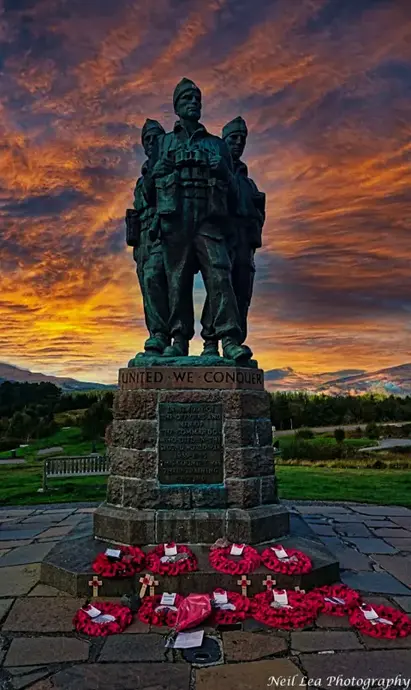 نیل لی مجسمه یادبود جنگی برنزی بر روی پایه سنگی با کلمات «متحد، پیروز میشویم» ایستاده است. پایه با تاجهای خشخاش قرمز و صلیبهای چوبی تزئین شده که نماد یادآوری و احترام است. پشت بنای یادبود، غروب دراماتیک آسمان را با سایههای نارنجی آتشین و بنفش عمیق پر میکند.