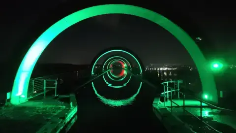 کوین هامیل مجموعهای از طاقهای سبز روشن، تونل اثیری بر فراز یک آبراه آرام ایجاد میکنند و بازتابهایشان دایرههای کامل تشکیل میدهند. سازه به طور دراماتیک در برابر آسمان شب تاریک روشن شده و نورهای شهر دور در پسزمینه به طور ضعیف دیده میشود. نردههای فلزی و سکوها پیشزمینه را قاب میکنند.