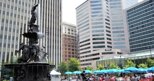 یک فواره بلند و تیره‌رنگ در سمت چپ، که توسط آسمان‌خراش‌های مرکز شهر سینسیناتی در پشت آن احاطه شده و در سمت راست ردیف‌های میزهای با چترهای سبز قرار دارند