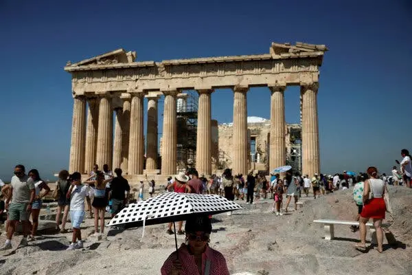 فردی چتری با طرح شطرنجی سیاه و سفید در دست دارد. در پسزمینه، پارتنون، سازه سنگی باستانی با ستونهایش، در میان جمعیت دیده میشود.