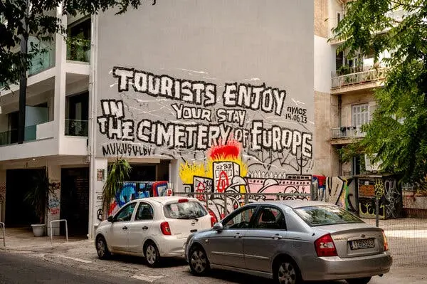 دیوار ساختمانی در شهر پوشیده از گرافیتی است، از جمله عبارت «توریستها، از اقامتتان در قبرستان اروپا لذت ببرید». دو خودرو در خیابان پارک شدهاند.