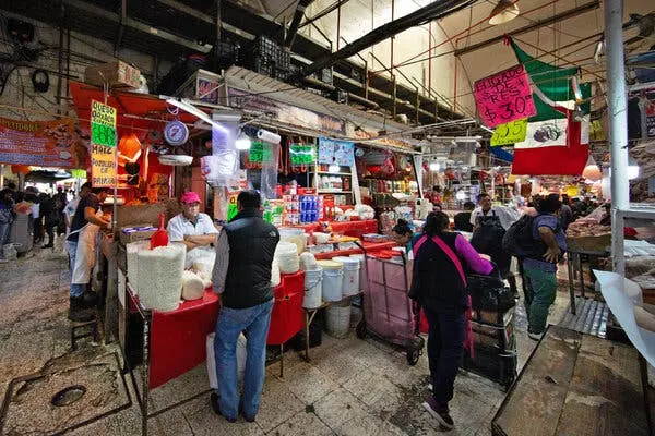 صحنهای از یک بازار شلوغ، با فروشندگانی که از ظروف بزرگ، سطلهای سفید و قفسههای پر از جنس، مواد غذایی میفروشند. پرچم بزرگ مکزیک با رنگهای قرمز، سفید و سبز بر فراز گذرگاهی شلوغ بین غرفهها آویزان است.