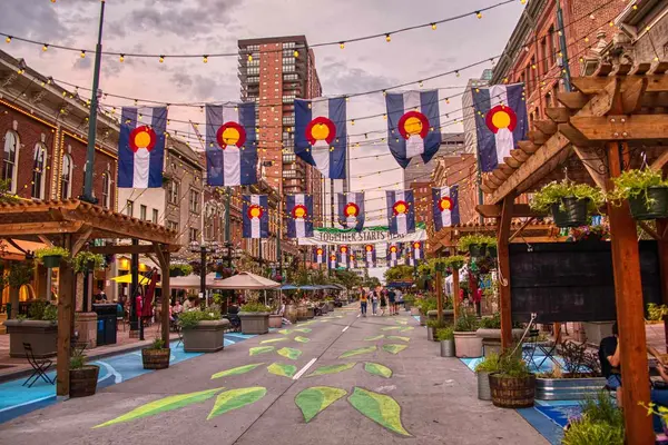 بازار خیابانی شهری در فضای باز با پرچمهای کلرادو، سبزینههای تزئینی و چراغهای رشتهای بالای سر